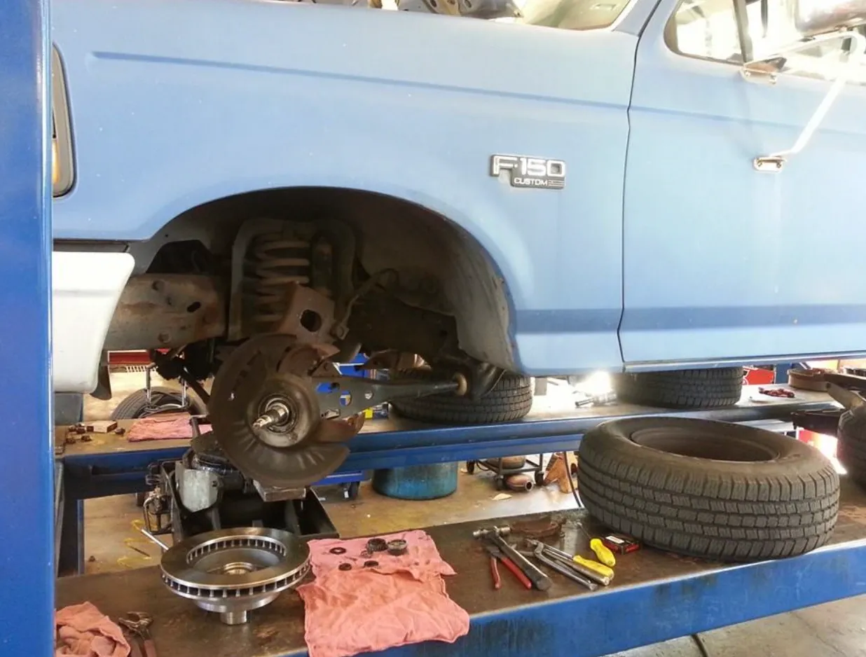 Screen Shot 2025-04-06 at 10.16.21 PM Blue Ford F-150 on a service lift with the rear wheel removed showing brake rotor and suspension components, spare tire and tools on the lift platform in a family-owned Tucker, GA auto repair shop.