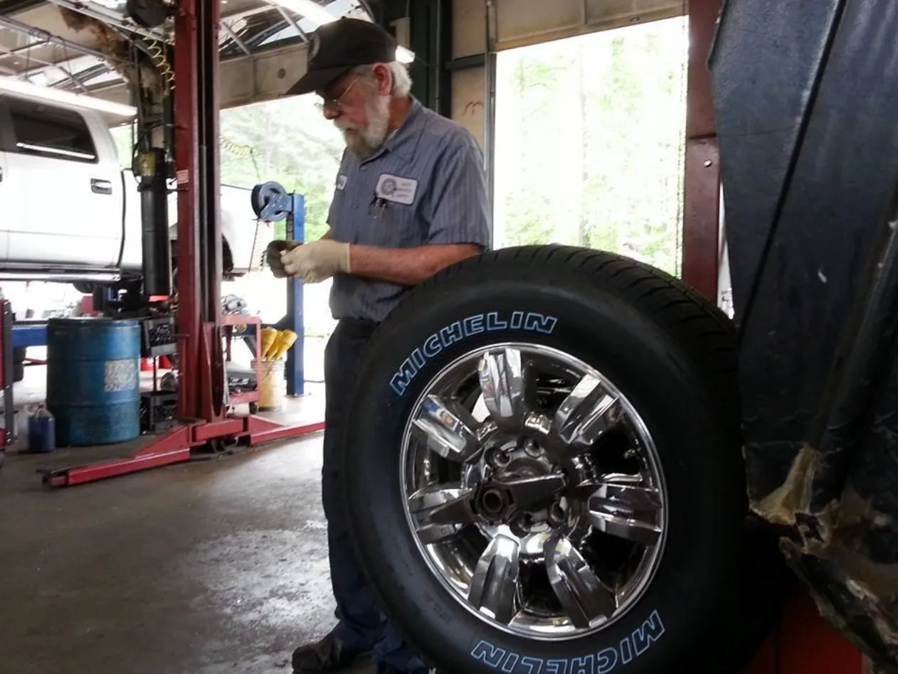 Screen Shot 2025-04-06 at 10.17.27 PM Mechanic in a family-owned Tucker, GA auto repair shop holding a tool next to a removed Michelin tire and shiny chrome wheel with a vehicle on a hydraulic lift in the background