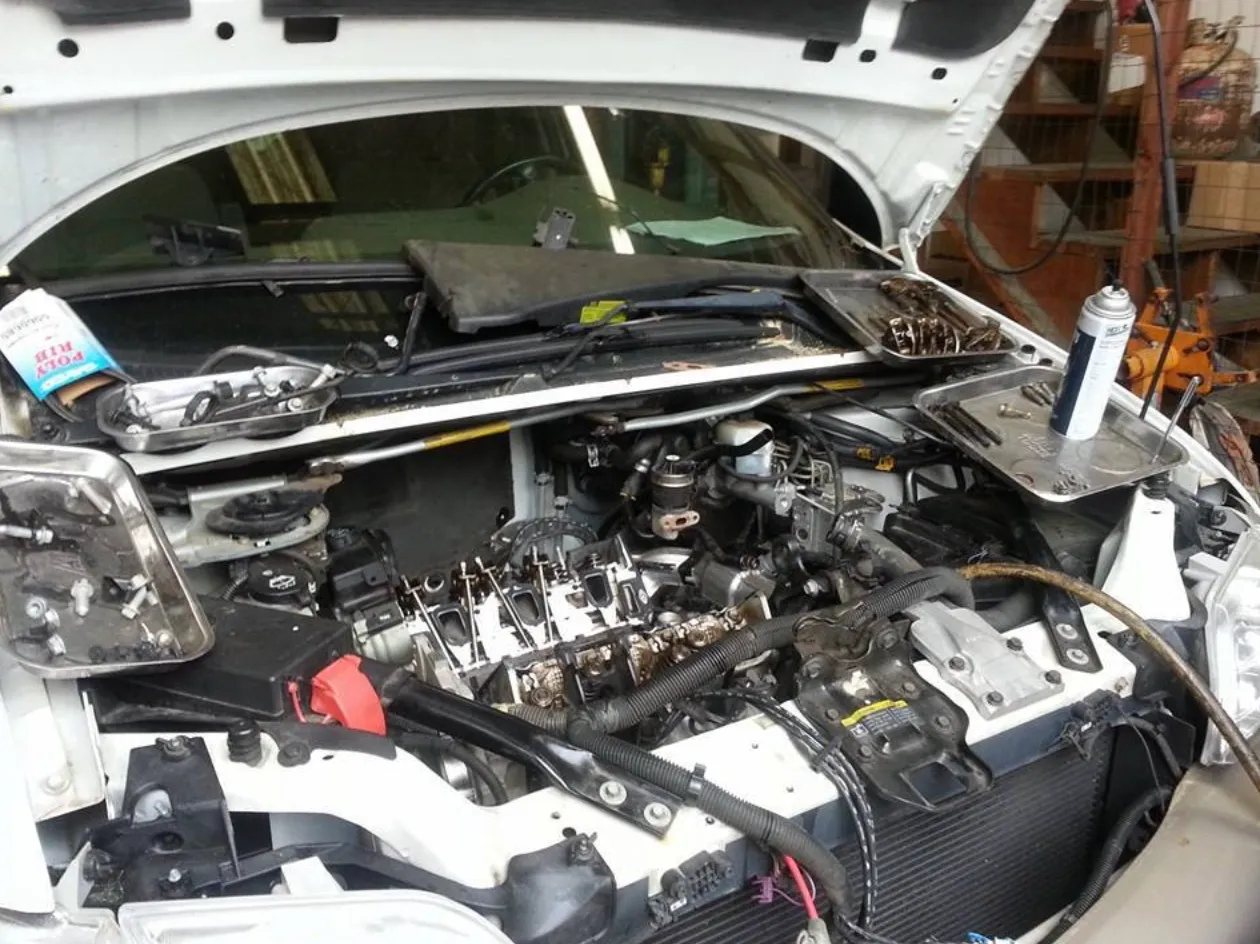 Screen Shot 2025-04-06 at 10.17.36 PM White car with its hood open showing a partially disassembled engine bay, exposed valve train and wiring with mechanic’s tools, spray can, and trays arranged on the fenders