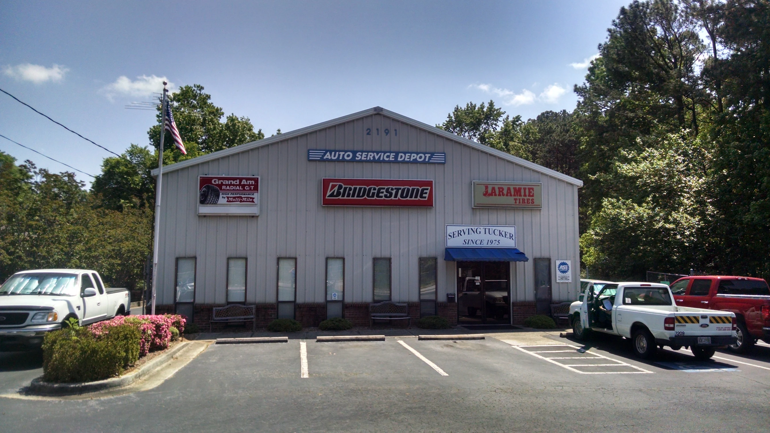 storefront Auto Service Depot metal shopfront with Bridgestone and Laramie Tires signs, blue awning reading "Serving Tucker since 1975", parked pickup trucks in front, American flag on a pole, trees surrounding the Tucker, GA auto repair shop
